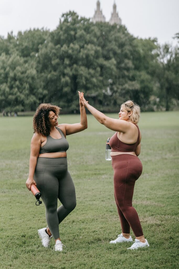 Two women in sportswear celebrate with a high five during a workout in a grassy park.